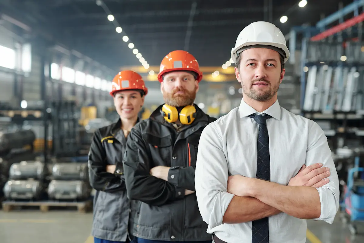 Three workers in safety gear stand inside an industrial warehouse. The man at the front wears a white hard hat and tie, while the two behind him wear orange hard hats and safety jackets.