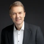 A middle-aged man in a dark suit and white shirt smiles against a dark background.
