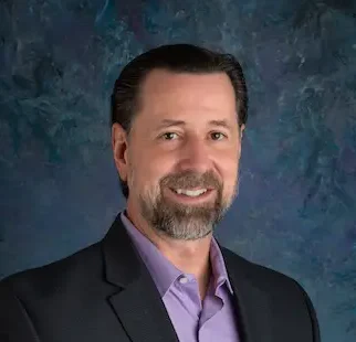 Man in a dark blazer and purple shirt smiles with hands in pockets against a textured blue background.