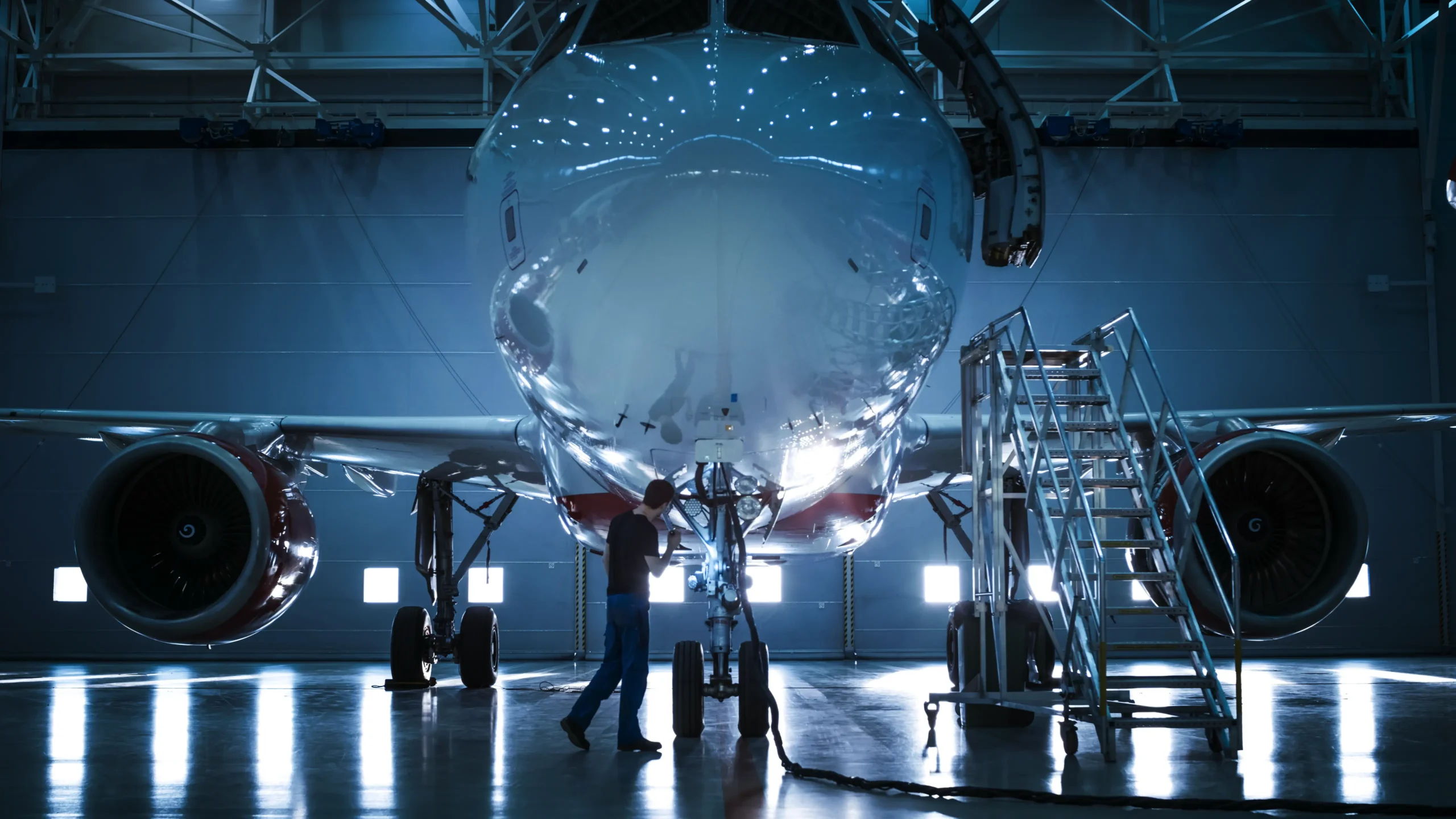 Technician inspecting the nose of a large airplane in a hangar, with a ladder nearby and light shining through windows.