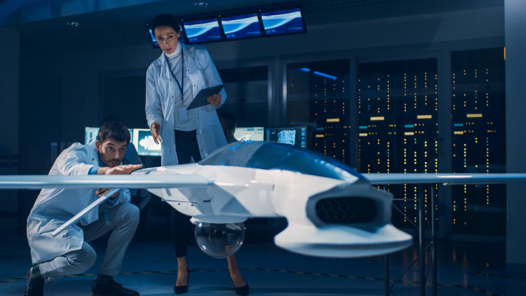 Two engineers in a lab coat and suit examine a model drone aircraft inside a technology lab. They are surrounded by computer servers and display screens.