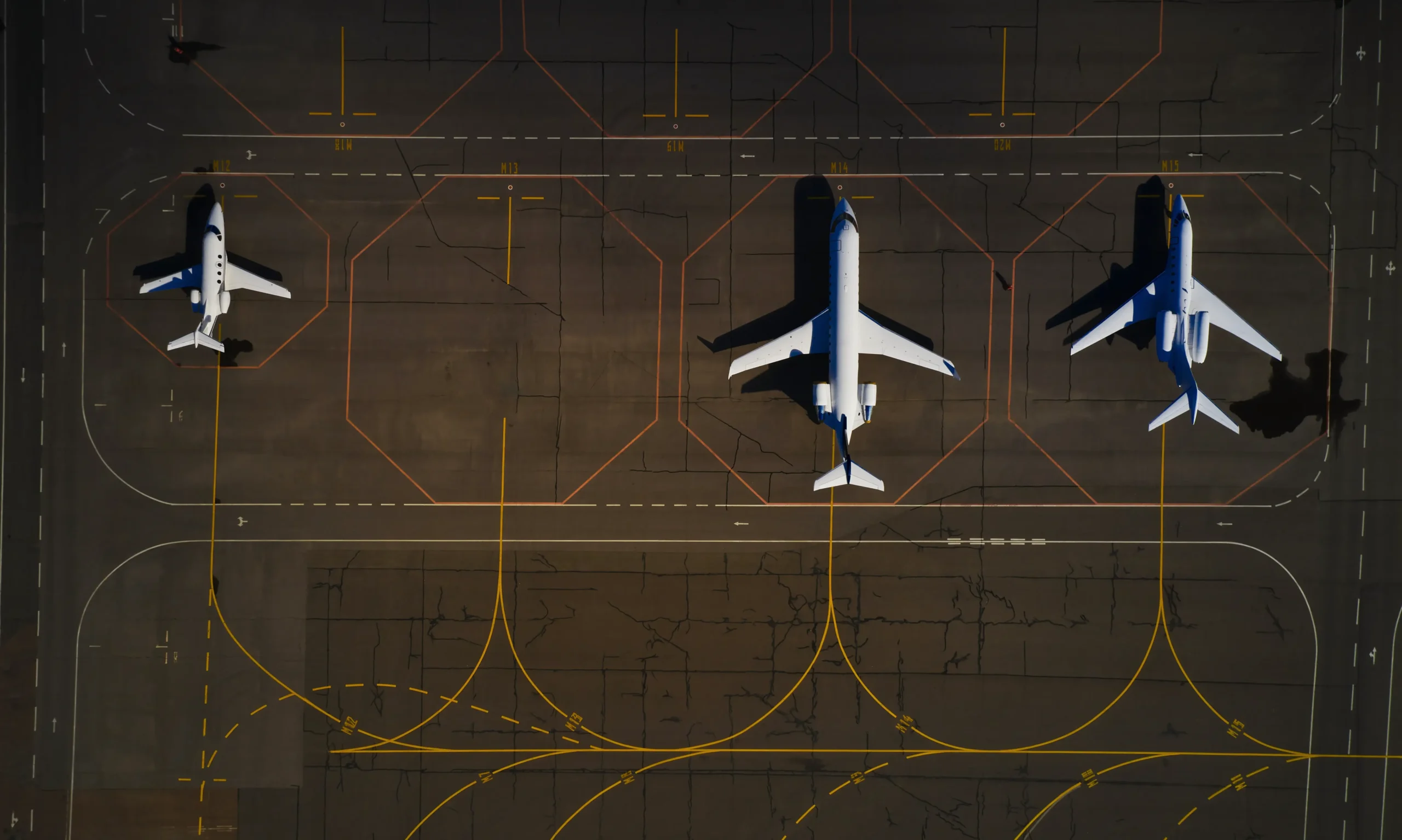 Aerial view of three airplanes parked on an airport tarmac, with distinct yellow lines and markings visible on the ground.