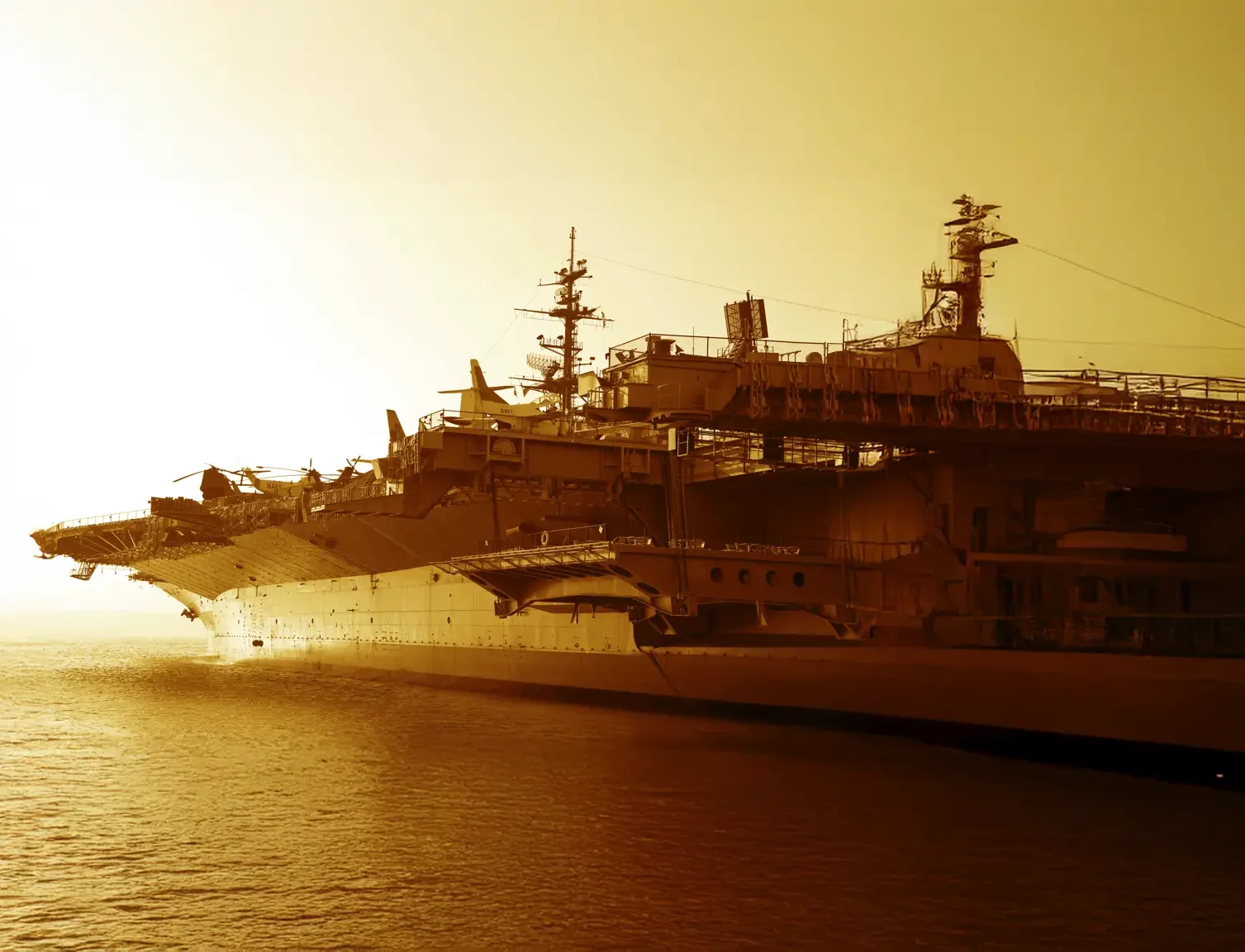 Large aircraft carrier docked at a harbor during sunset, with a golden hue illuminating the scene and calm water surrounding the vessel.