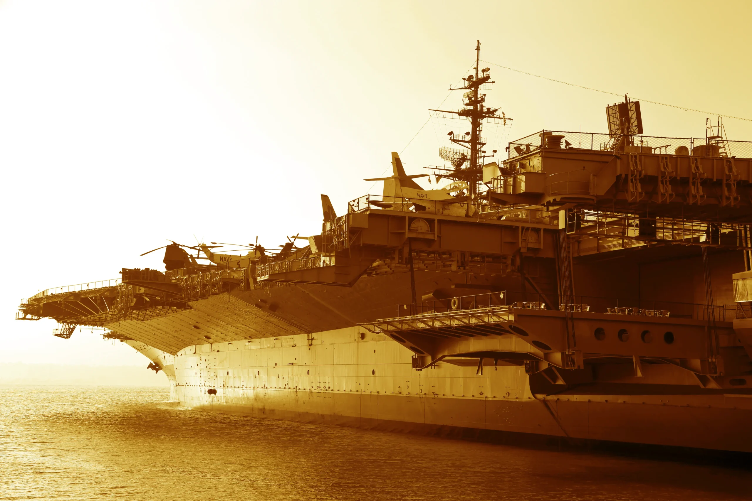 An aircraft carrier is stationed in calm waters under a warm, golden sunset, with helicopters visible on its deck.