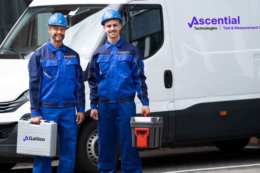 Two technicians in blue uniforms and helmets stand in front of a white van, holding equipment cases labeled "Galileo" and "Ascential Technologies | Test & Measurement.
